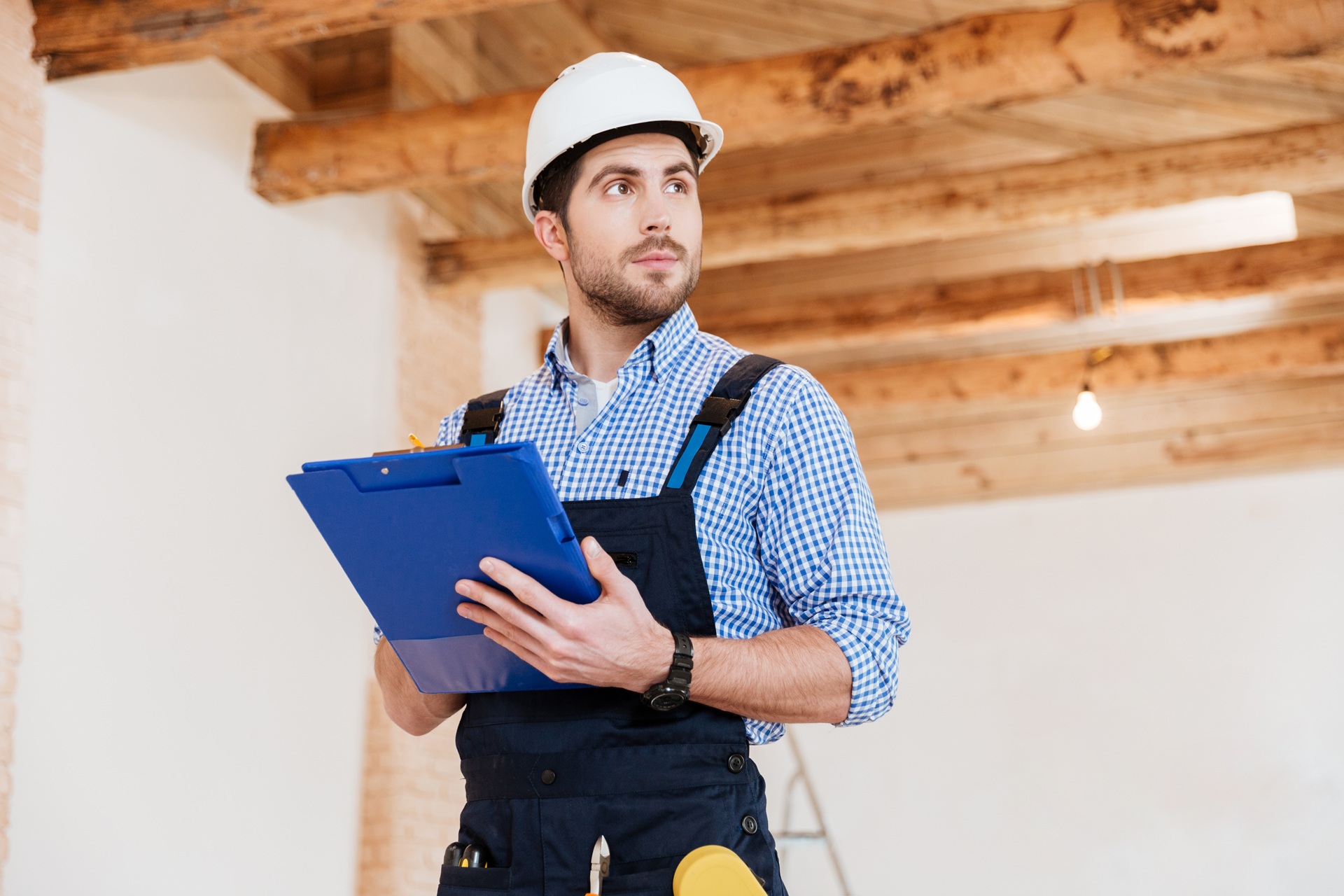 Pensive builder in hardhat with clipboard and pencil indoors